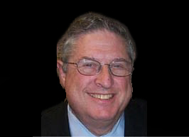 Hal Bookbinder, Wearing Glasses And A Suit, Smiles In A Formal Close-Up Portrait Against A Dark Background