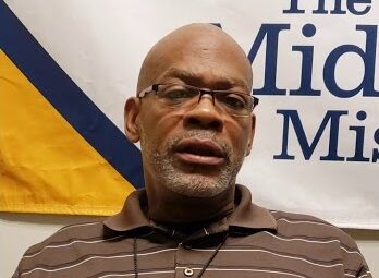 Man Wearing Glasses And A Brown Striped Polo Shirt Speaking During An Interview With The Midnight Mission Banner In The Background