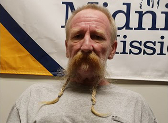Max With A Long Braided Mustache And Gray T-Shirt Standing In Front Of The Midnight Mission Banner, Portrait Photo