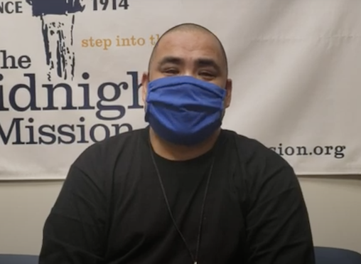 Man In A Blue Face Mask And Black Shirt Sitting In Front Of The Midnight Mission Banner, Sharing His Testimonial Story