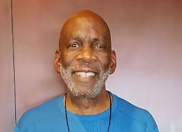 Bruce Smiling Warmly In A Blue Sweater During A Headshot Against A Brown Background, Showing A Friendly And Approachable Expression