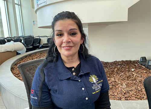Frankie Smiles While Seated Indoors Wearing A Midnight Mission Polo Shirt In A Bright Lobby Area With Chairs Behind Her