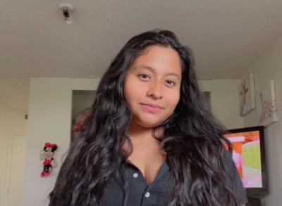 Woman With Long Dark Wavy Hair Wearing A Black Shirt, Smiling Gently In A Home Setting With Wall Decor Visible