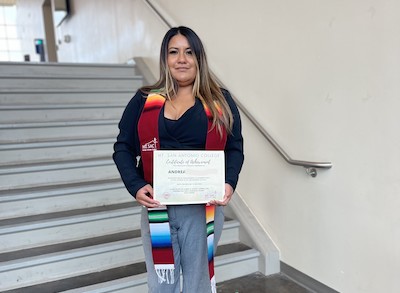 Andrea M. Stands On A Staircase Holding A Certificate And Smiling While Wearing A Colorful Graduation Stole