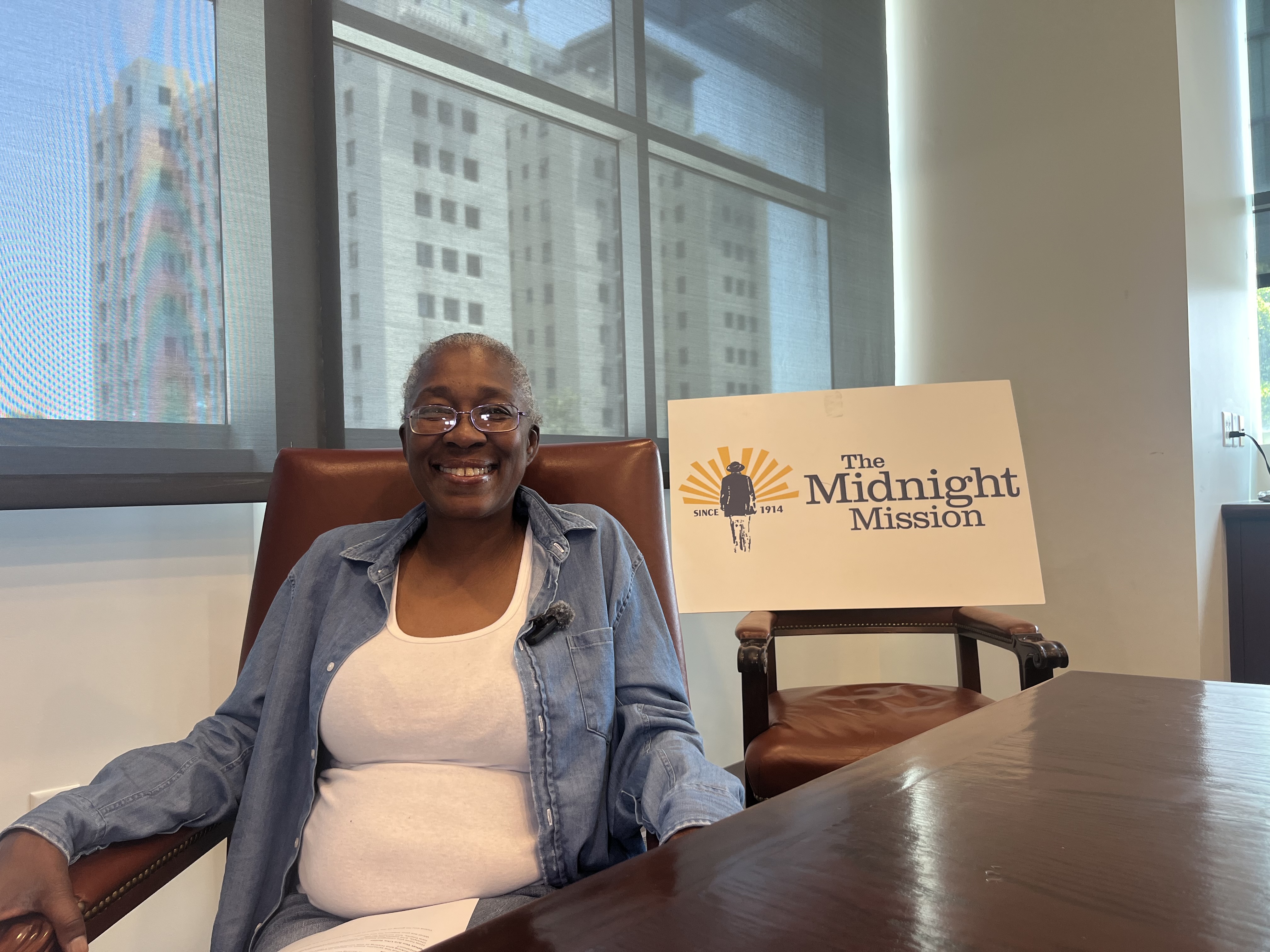 Smiling Woman Seated At Desk, Having White Board Written Midnight Mission Beside, Sharing Her Story In An Office Interview Setting