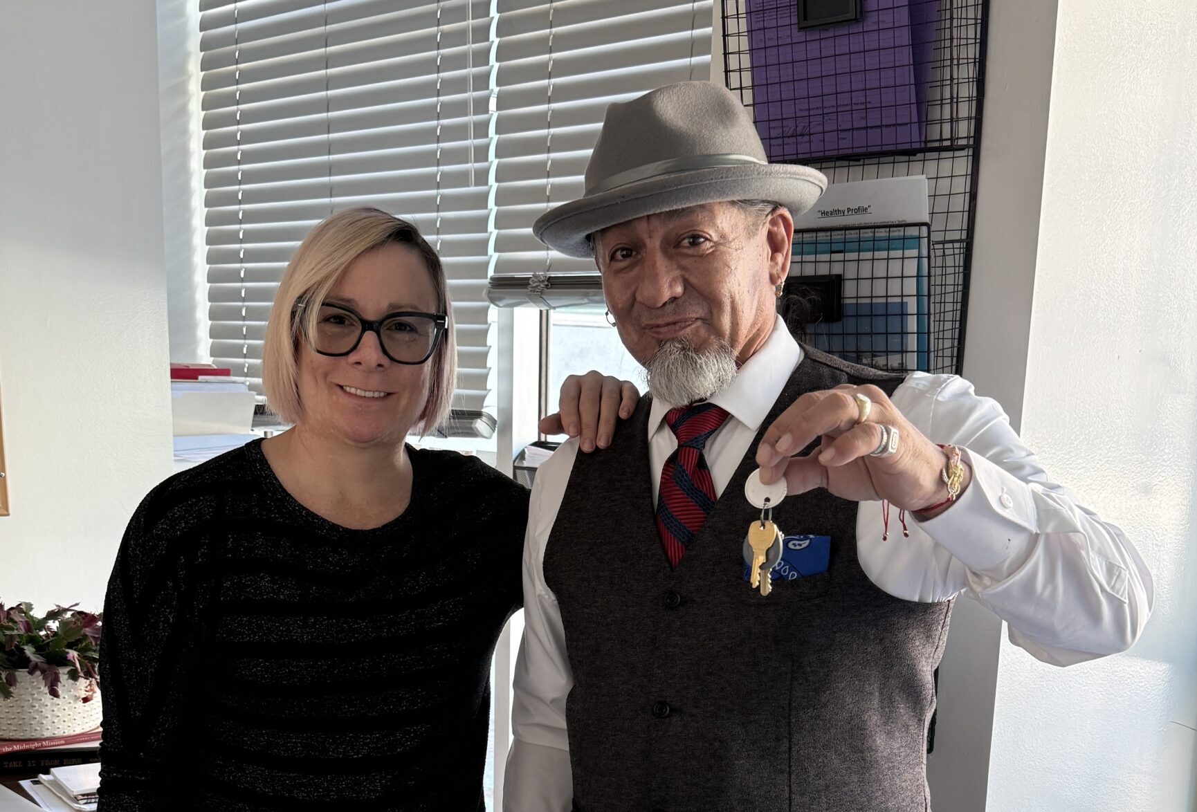 A Man Holds Up A Set Of Keys While Standing Beside A Smiling Woman Indoors, Both Posing For The Camera