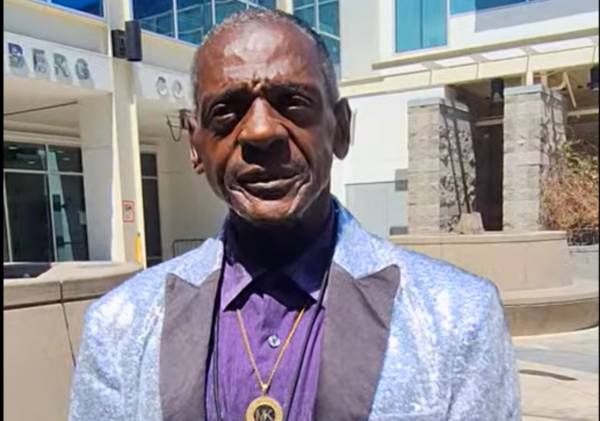 Man Wearing A Shimmering Silver Blazer Over A Purple Shirt, Standing Outdoors With A Confident Smile In Front Of A Modern Building