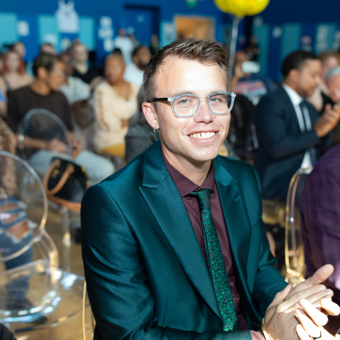 Kyle Meek Smiles In A Teal Suit While Attending A Midnight Mission Gathering, Seated Among Guests During The Event