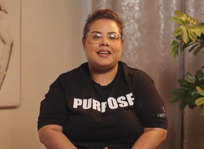 Jubiline Wearing Glasses And A Black Purpose T-Shirt, Speaking Indoors Near A Green Plant, Sharing Her Personal Story And Journey Today