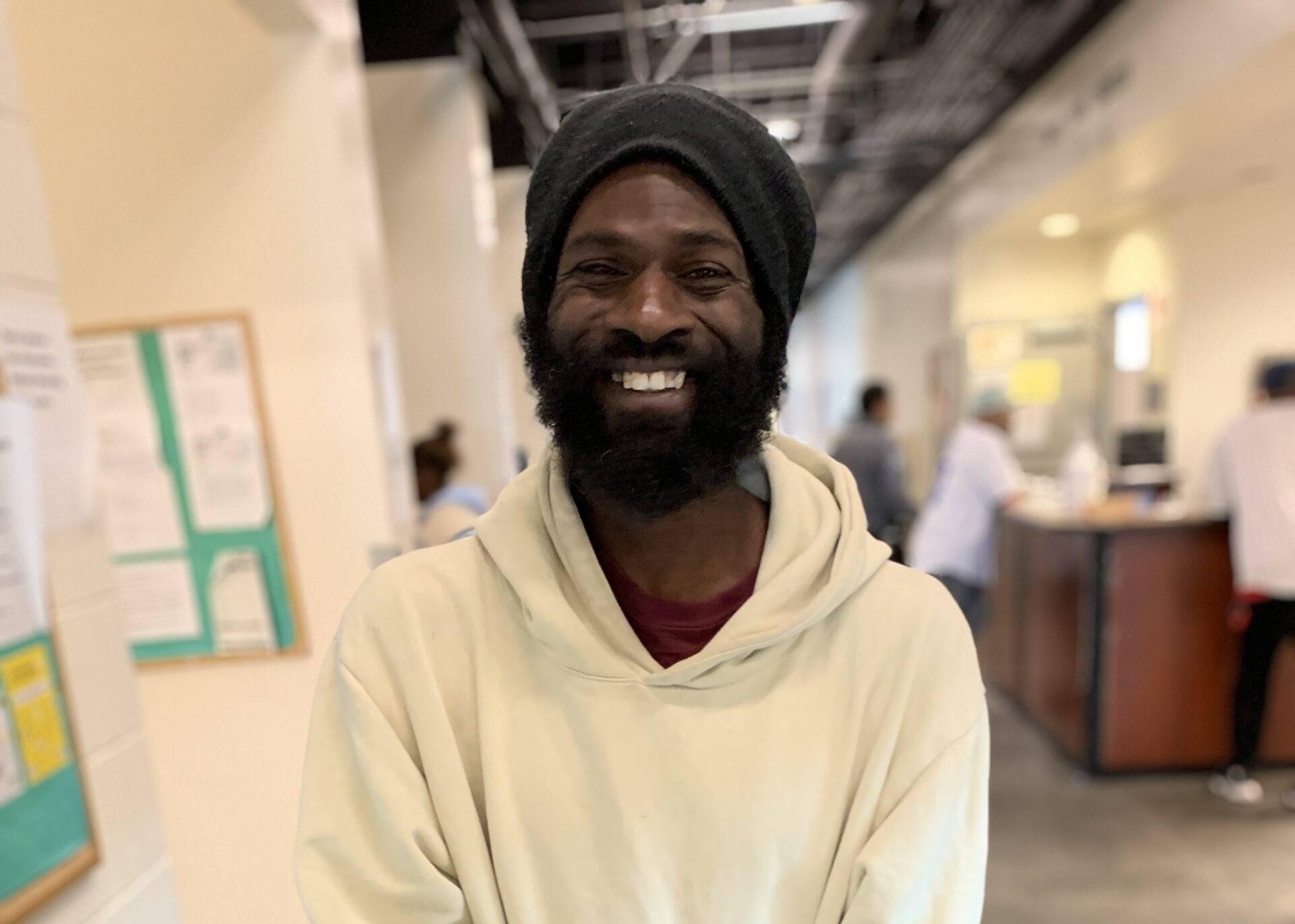 Terrance Smiling With A Full Beard, Wearing A Black Knit Cap And Cream Hoodie Inside The Facility With A Welcoming Expression