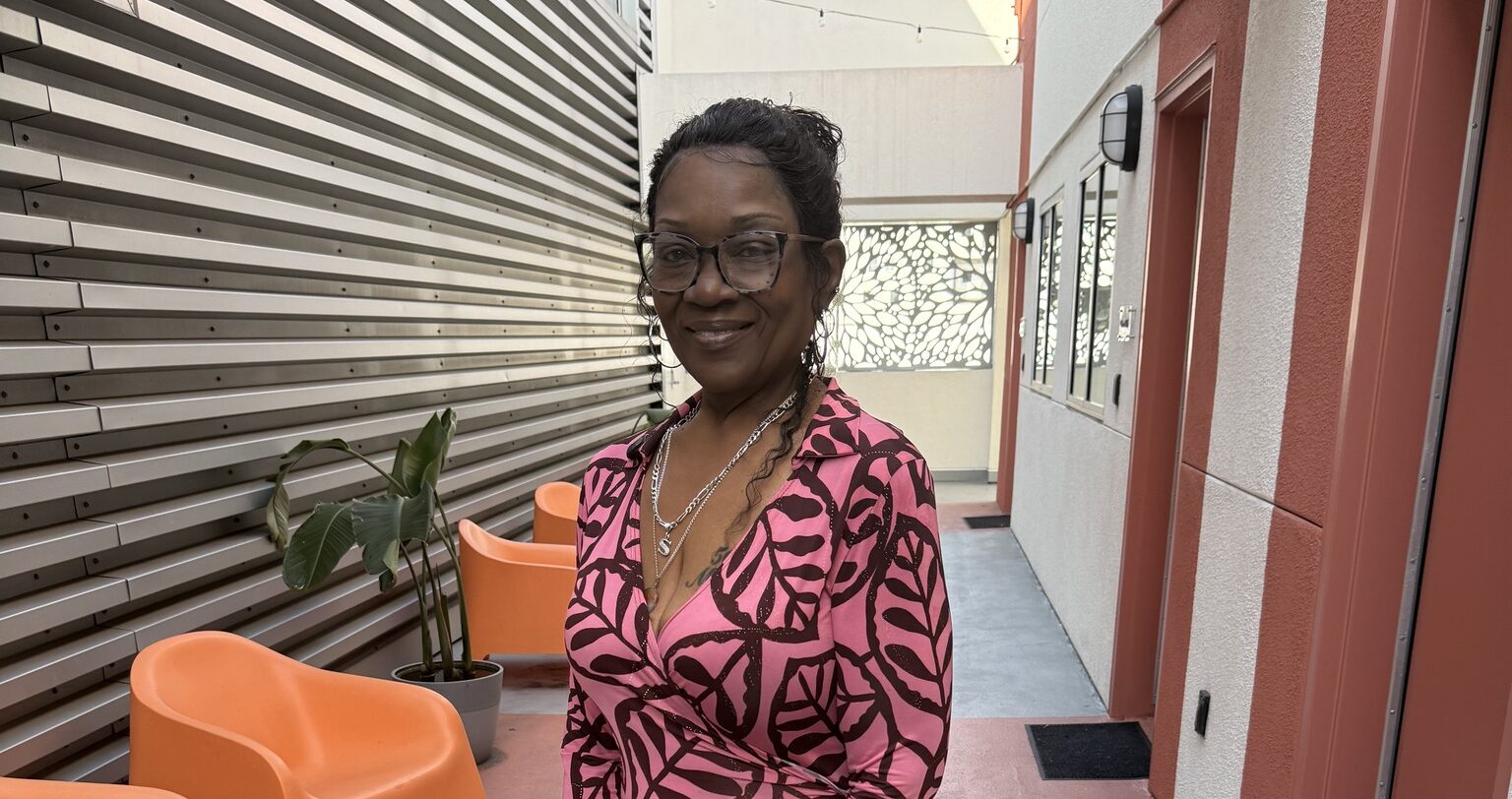 Stacey, Wearing Glasses And A Pink Patterned Top, Smiles In An Outdoor Courtyard With Decorative Metal Paneling