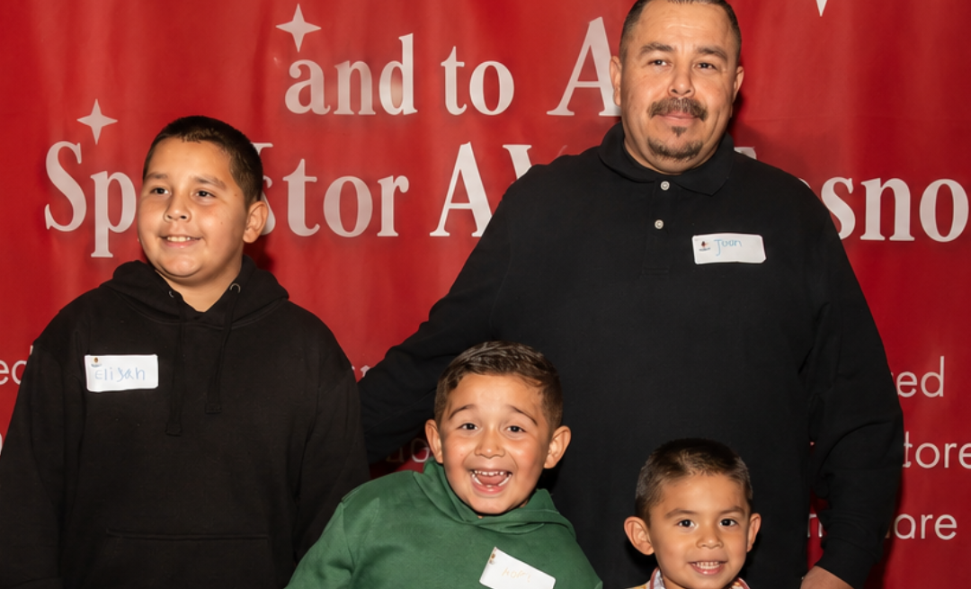 Juan And His Three Smiling Sons Wearing Name Tags, Posing Together At A Holiday Event With A Red Festive Backdrop